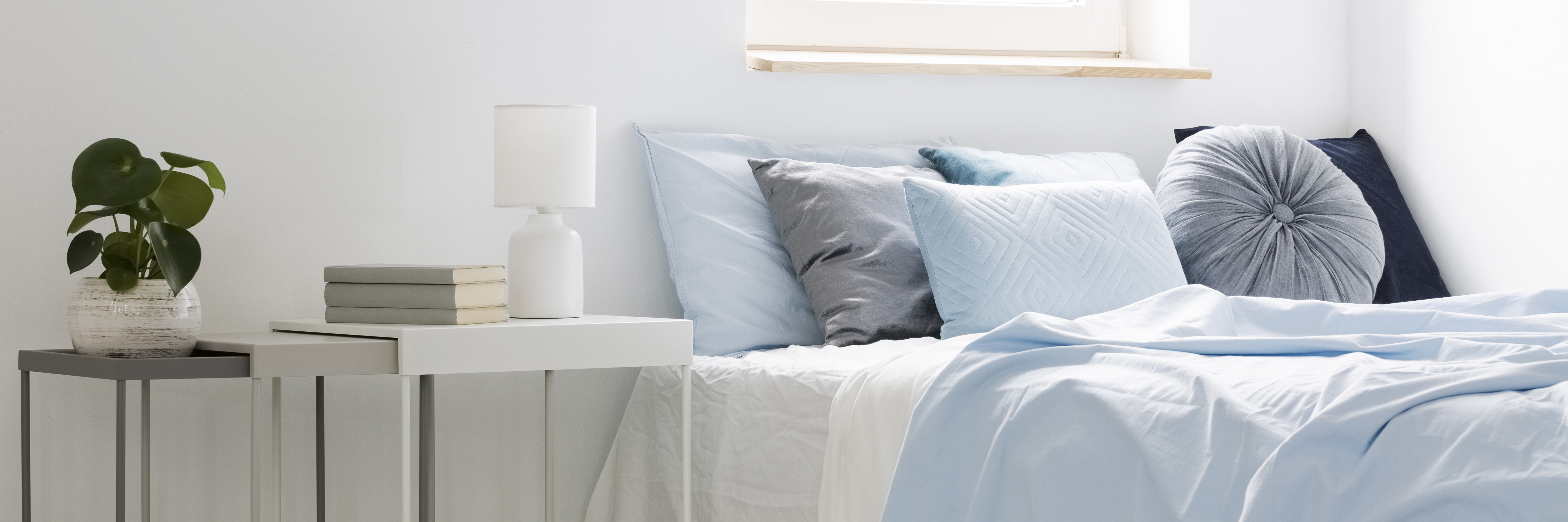 bed-with-blue-bedding-and-cushions-standing-next-to-white-tables-with-books-lamp-and-plant-in-bedroom-interior